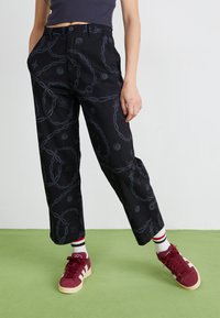 Pantaloni neri con un motivo a fantasia che presenta catene grigie e motivi circolari. Hanno un taglio dritto e sono abbinati a sneakers bordeaux.