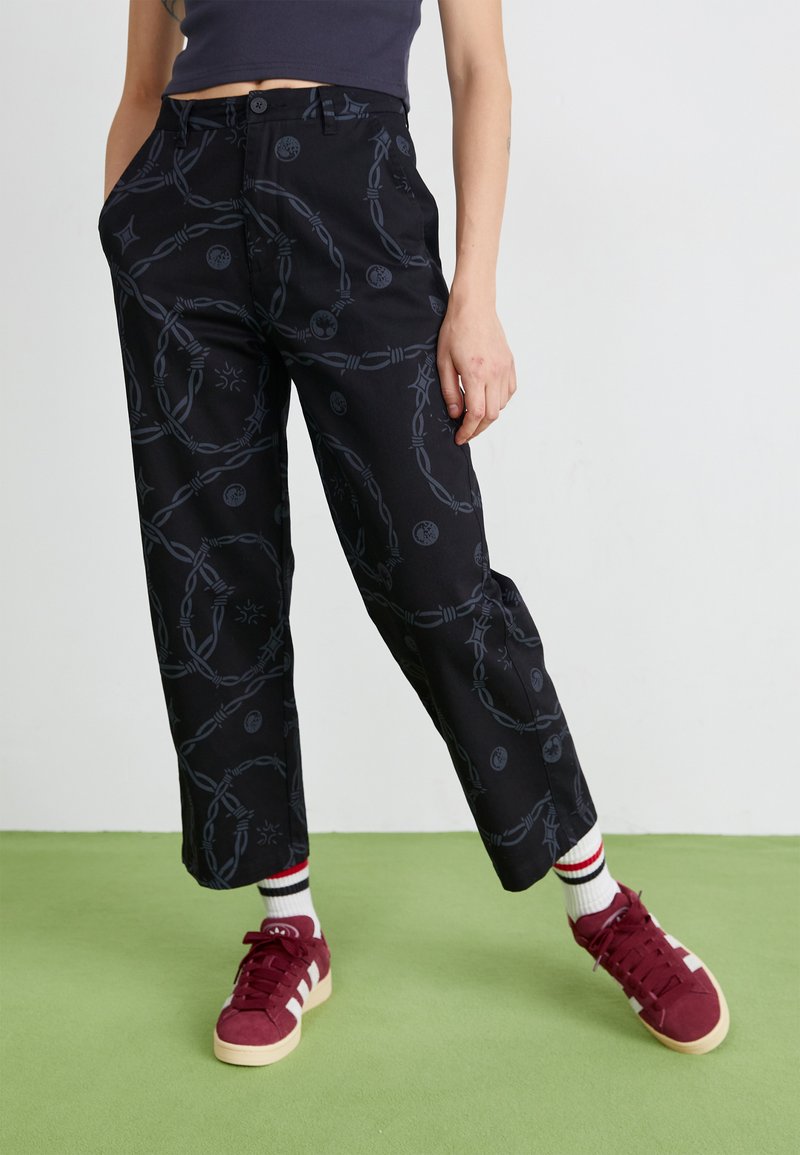 Pantaloni neri con un motivo a fantasia che presenta catene grigie e motivi circolari. Hanno un taglio dritto e sono abbinati a sneakers bordeaux.