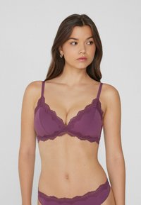 Jeune femme aux longs cheveux bruns portant un ensemble de lingerie violet à bord dentelé, regardant vers sa droite devant un fond uni.