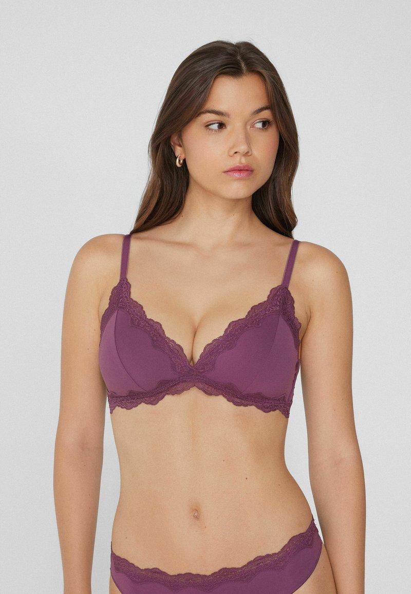 Jeune femme aux longs cheveux bruns portant un ensemble de lingerie violet à bord dentelé, regardant vers sa droite devant un fond uni.