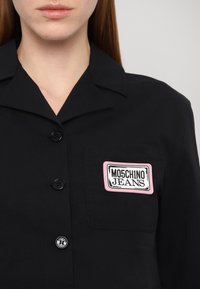 Musta särg, millel on krae, millel on roosa ja valge logo märk pealkirjaga "MOSCHINO JEANS" ning kaks musta nupukest ees.