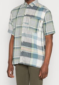 Camicia a maniche corte in cotone con un motivo a quadri multicolore, dotata di chiusura con bottoni e tasca sul petto, prevalentemente in tonalità blu e verde.