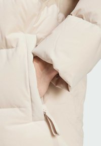 Beige puffer jacket detail with a visible pocket, featuring a textured fabric and secure closure toggle, highlighting its soft material.