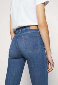 Pantalones vaqueros de mezclilla azul con una cintura alta, que presentan un parche de cuero marrón, bolsillos traseros y un diseño ajustado. Textura suave.