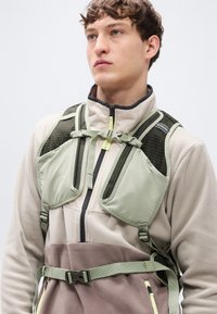 Ανοιχτό πράσινο ζεύγμα με επενδεδυμένους ιμάντες ώμου, ρυθμιζόμενες πόρπες και υφασμάτινα πάνελ. Φοριέται πάνω από ένα μπεζ και καφέ fleece μπουφάν.