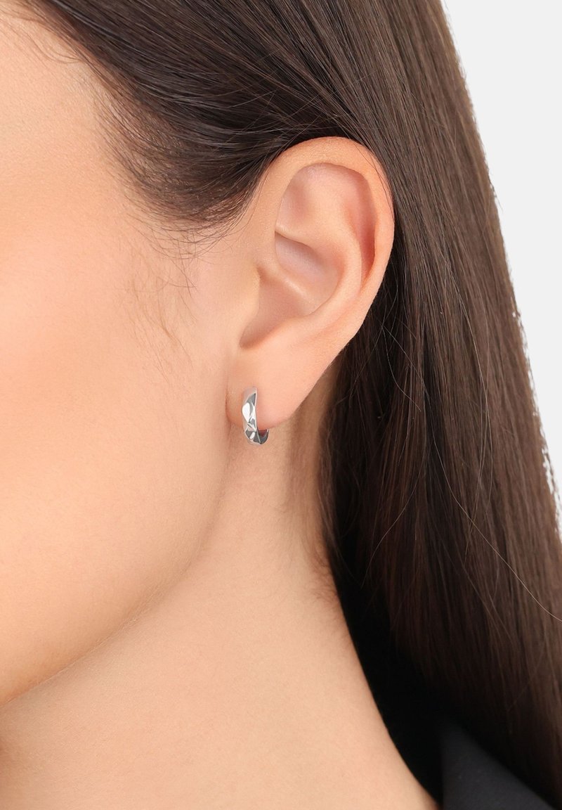 Una piccola orecchino a cerchio in argento intrecciato indossato all'orecchio sinistro di una donna con i capelli castani lisci portati dietro l'orecchio.