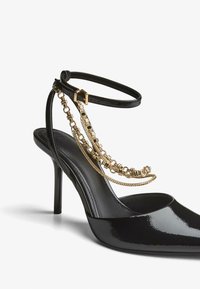 Črne lakaste usnjene čevlje s peto in izostrenim nosom, z zapenjanjem okoli gležnja, ki vsebuje zlate verižne detajle za dodano stil.