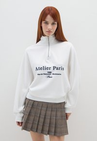 Felpa bianca con zip con "Atelier Paris" ricamato in blu navy; abbinata a una gonna a pieghe scozzese marrone con più pieghe.