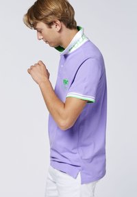 Lila Poloshirt aus Baumwolle mit weißem Kragen, der grünen "POLO"-Aufschrift. Grüne und weiße gestreifte Akzente an den Ärmeln mit gesticktem Logo.