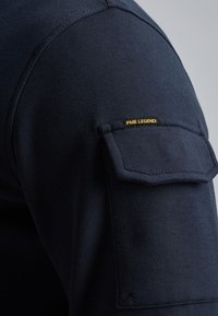 Camisa de manga larga de color azul marino con un tejido texturizado, que cuenta con un bolsillo con solapa en la parte superior del brazo y una etiqueta dorada de "PME LEGEND".
