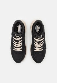 Baskets noires en tissu et daim, présentant un design texturé, des lacets crème et une semelle extérieure beige. Logo "MK" sur la languette.