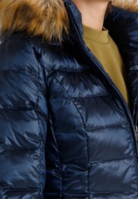 Laivastonsininen untuvatakki, jossa on sileä ja kiiltävä pinta, irrotettava karvavuorinen huppu sekä vetoketjullinen suljenta. Hienovarainen ommeltu paneelikuva.