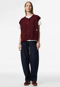 Vinröd oversized väst med texturerad fleecefinish, rund halsringning och knäppning framtill, lager-på-lager över en vit T-shirt. Mörka denimbyxor.