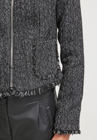 Gros plan sur une veste en tweed texturée noir et blanc avec une fermeture éclair, des bords effilochés et une poche patch sur un pantalon en cuir noir.