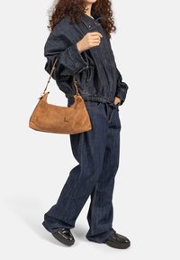 Hellbraune Wildlederhandtasche mit geschwungener Form und kurzem Trageriemen, kombiniert mit einem lockeren, dunklen Denim-Outfit mit betonter Taille.