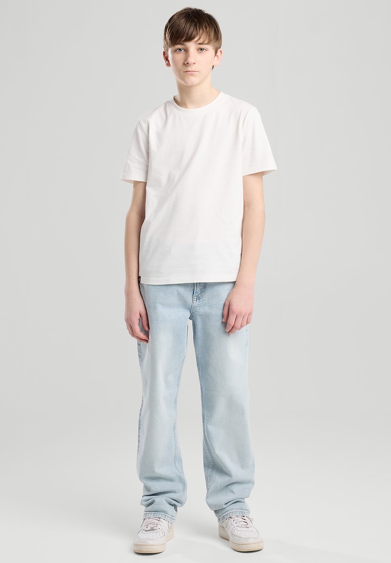 Adolescent debout, portant un t-shirt blanc uni, un jean bleu clair et des baskets blanches, sur un fond gris clair uni.