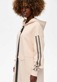 Vrouw met krullend haar die een beige jas met capuchon draagt, met een zwarte rits en dubbele zwart-witte streepdetails op de mouwen.