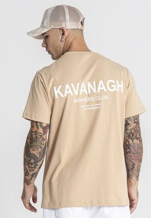 Uomo con t-shirt beige "KAVANAGH WINNERS CLUB" e cappellino beige a rete, con braccia tatuate, che guarda di lato su uno sfondo chiaro.