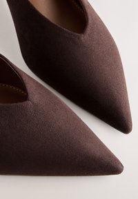 Paire de chaussures pointues en daim brun foncé avec empeigne en forme de V, vues en gros plan sur un fond clair.