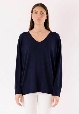 Donna con lunghi capelli scuri che indossa un maglione scollo a V blu navy ampio e pantaloni bianchi, in piedi davanti a uno sfondo chiaro uniforme.