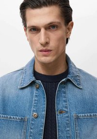 Jachetă din denim de culoare albastru deschis, cu guler structurat, închidere cu nasturi în față și un buzunar, purtată peste un pulover ribbit de culoare albastru închis.