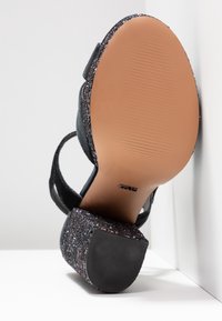 Sandale à talons pailletée noire avec une semelle texturée, caractérisée par une plate-forme épaisse ornée et de fines sangles de cheville. La semelle est lisse avec des nervures.