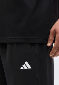 Gros plan d'une personne portant un pantalon de sport noir avec un logo blanc et un t-shirt noir, montrant le bas du torse et la main droite.