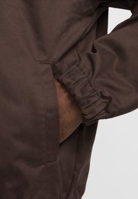 Hnědá bunda s texturovanou, odolnou tkaninou, s elastickými manžetami a částečně viditelnou boční kapsou.
