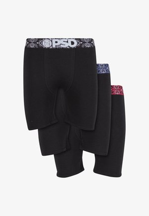 Mężczyzna bez koszulki z piegami, noszący czarne bokserki PSD z czarno-białym pasem, stojący boso na białym tle.