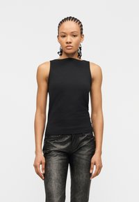 Femme aux cheveux tressés portant un haut noir sans manches et un pantalon en cuir noir texturé, debout devant un fond clair uni.