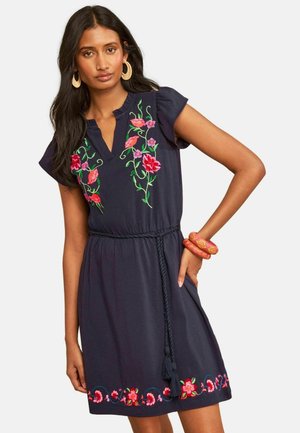Frau trägt ein dunkelblaues Kleid mit bunter Blumenstickerei, kurzen Ärmeln, geflochtenem Taillenband und orangen Armreifen am rechten Handgelenk.