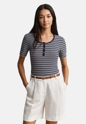 Nő tengerészkék-fehér csíkos rövid ujjú ingben, gombos gallérral, magas derekú fehér sortba tűrve, barna övvel.