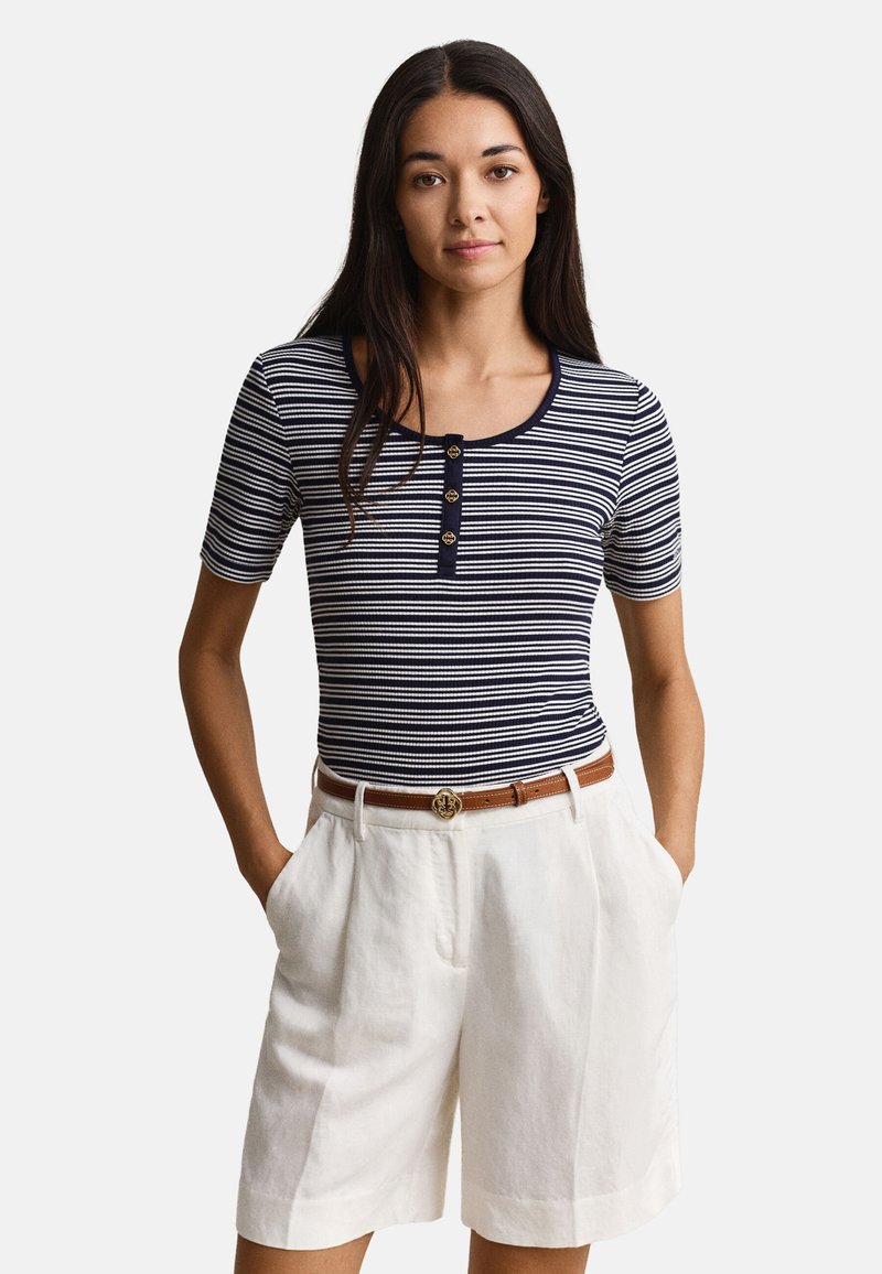 Γυναίκα με μπλε ναυτικό και λευκό ριγέ κοντομάνικο πουκάμισο με κουμπιά, μπαίνει μέσα σε λευκές σόρτς με ψηλή μέση και καφέ ζώνη.