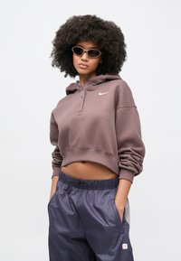 Γυναίκα με κατσαρά μαλλιά που φορά γυαλιά ηλίου cat-eye, μοβ cropped hoodie με το λογότυπο της Nike και μπλε αθλητικό παντελόνι, με τα χέρια στις τσέπες.