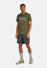 Olivgrön t-shirt med en berggrafik, kombinerad med mörkgrå shorts. Modellen har på sig ljusgrå strumpor och tan-färgade löparskor.