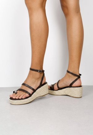 Piernas femeninas con sandalias cuña de cuero marrón con correa en el tobillo y suelas de plataforma de yute trenzado sobre un suelo gris y fondo blanco.