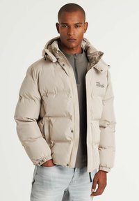 Lengvas bešviesos puffer parkas, šviesiai smėlinės spalvos, su užtrauktuku priekyje, elastingomis rankovėmis ir kapišonu. Sklandži tekstūra su subtiliu ženklu ant krūtinės.