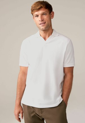 Cămașă polo albă, cu o textură moale, mâneci scurte și guler. Modelul poartă pantaloni maro închis și stă în fața unui fundal neutru.