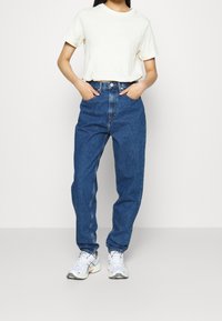 Světle béžové bavlněné tričko, modré džíny s vysokým pasem, volný střih a bílé sportovní boty. Jednoduchý design, žádné viditelné vzory nebo loga.