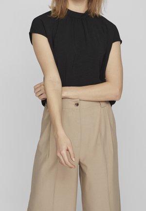 Frau trägt ein schwarzes kurzärmeliges Oberteil und beige, hoch taillierte weite Hosen mit Knopfverschluss, steht mit verschränkten Armen.