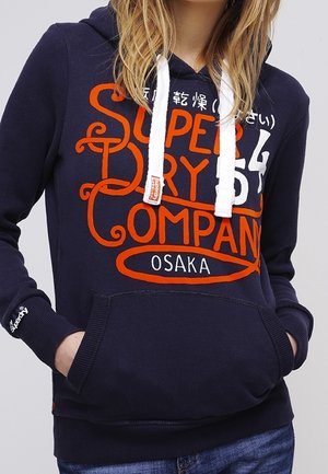 Kobieta ubrana w granatową bluzę z kapturem z dużym pomarańczowo-białym napisem "Super Dry Company Osaka", białymi sznurkami i rękami w przedniej kieszeni.