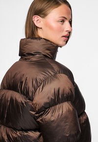 Brun puffjacka med hög krage, tillverkad av glansigt, quiltat material. Har en rundad form och tydliga plisserade partier.