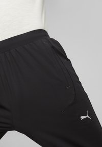 Čierne športové nohavice vyrobené z jemného, ľahkého materiálu. Obsahujú nastaviteľný pás so šnúrkou, bočné vrecká a malé biele logo Puma na dolnej časti nohy.