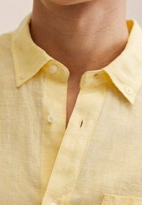 Chemise en lin jaune avec un col, deux boutons de fermeture et une poche poitrine. Présente un tissu texturé avec des coutures visibles et un petit défaut.