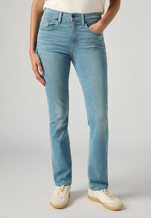 Personne portant un jean droit bleu clair et des baskets écru, debout, une main dans la poche et l'autre main détendue sur le côté.
