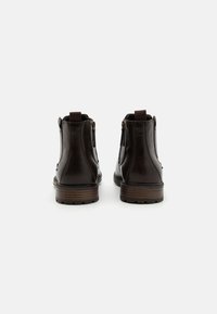 Botas de cuero marrón oscuro hasta el tobillo, con cremalleras laterales, suelas de goma y lengüetas en la parte trasera. Textura lisa y detalle de costura en contraste.