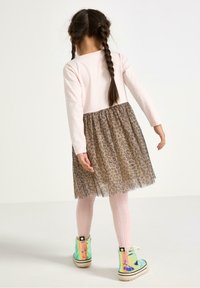 Maglietta rosa a maniche lunghe con una gonna in tulle a stampa leopardata marrone. L'outfit comprende delle calze rosa pastello e degli stivali lucidi iridescenti con lacci.