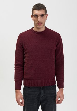 Burgundy knitted sweater with a round neckline, ribbed cuffs and hem, textured fabric, and a small tag at the side seam.