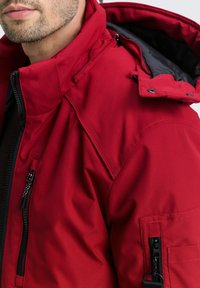 Raudona, izoliuota striukė su aukštu apvadu, užtrauktukais užsegamomis kišenėmis ir nuimamu gobtuvu. Ypatybės: tekstūruota medžiaga ir juodos detalės.