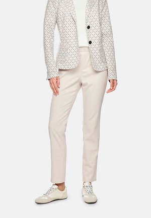 Frau trägt hellbeigen gemusterten Blazer, weißes Top, beige gerade Hose und cremefarbene Sneakers, steht mit entspannten Händen.